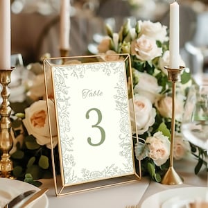 May include: A table number card with a gold frame, featuring the number 3 in green. The card is surrounded by a floral design and the word "Table" is at the top. The background includes candles, flowers, and a table setting.