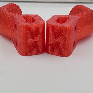 Puede incluir: Dos objetos rojos impresos en 3D con una forma curva y una base cuadrada. Cada objeto tiene un agujero rectangular en el centro de la base cuadrada y una serie de peque&ntilde;os agujeros en los lados de la base cuadrada.