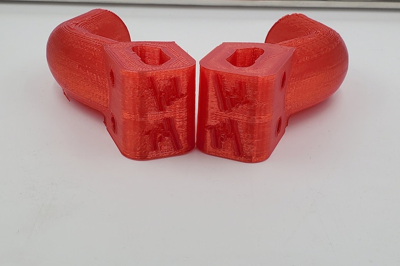 Puede incluir: Dos objetos rojos impresos en 3D con una forma curva y una base cuadrada. Cada objeto tiene un agujero rectangular en el centro de la base cuadrada y una serie de peque&ntilde;os agujeros en los lados de la base cuadrada.