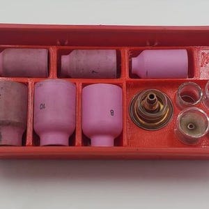 以下が含まれることがあります： 溶接トーチ部品が入った赤いプラスチック製のオーガナイザー。トレイにはピンク色のセラミックノズル、透明なガラスカップ、真鍮色のガスレンズが入っています。一部のノズルには番号が記載されています。