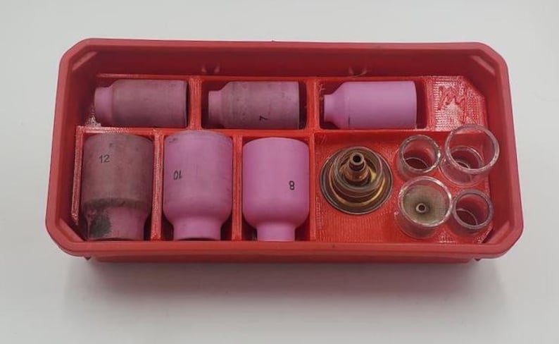 以下が含まれることがあります： 溶接トーチ部品が入った赤いプラスチック製のオーガナイザー。トレイにはピンク色のセラミックノズル、透明なガラスカップ、真鍮色のガスレンズが入っています。一部のノズルには番号が記載されています。