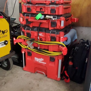 Puede incluir: Una pila de cajas de herramientas rojas con asas negras y una manguera amarilla y negra. La caja de herramientas inferior est&aacute; etiquetada como "Milwaukee Packout".