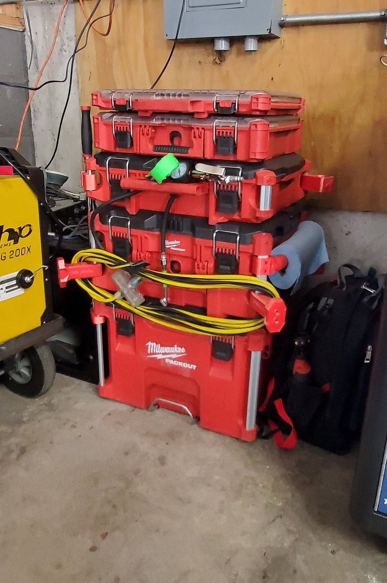 Puede incluir: Una pila de cajas de herramientas rojas con asas negras y una manguera amarilla y negra. La caja de herramientas inferior est&aacute; etiquetada como "Milwaukee Packout".