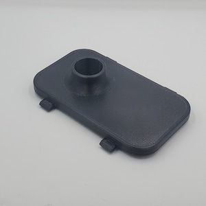 Peut inclure: Un objet rectangulaire en plastique noir imprimé en 3D avec un trou rond au centre. L'objet a quatre petites pattes sur le dessous.