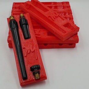 Op de afbeelding: Twee zwarte lasbrandermondstukken worden bewaard in een rode 3D-geprinte houder. De houder heeft de tekst "TMJ" erop gedrukt.