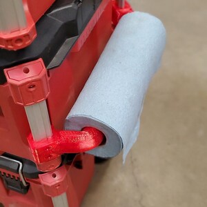 Puede incluir: Una caja de herramientas roja con un soporte para papel de cocina impreso en 3D fijado al lateral. El soporte es rojo y tiene un rollo de papel de cocina azul sujeto a &eacute;l.