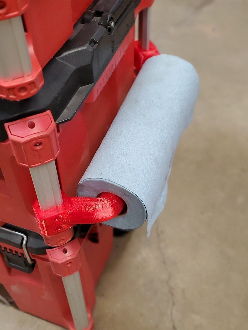 Puede incluir: Una caja de herramientas roja con un soporte para papel de cocina impreso en 3D fijado al lateral. El soporte es rojo y tiene un rollo de papel de cocina azul sujeto a &eacute;l.