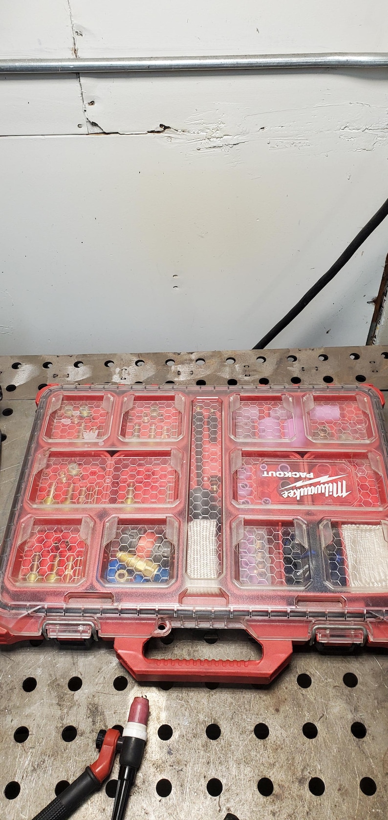 以下が含まれることがあります： 赤と透明なMilwaukee Packoutオーガナイザーケースには、さまざまなハードウェアとツールが詰まっています。 ケースは開いており、さまざまなサイズの継手とコンポーネントを備えた複数のコンパートメントが表示されます。 溶接トーチが前景にあります。