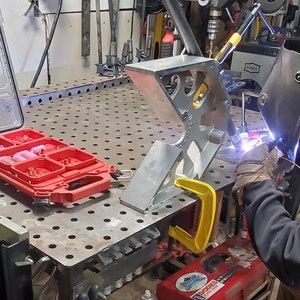 Może przedstawiać: Spawacz w czarnym swetrze z kapturem i masce spawalniczej pracuje nad metalowym projektem. Projekt jest zamocowany do metalowego stołu warsztatowego za pomocą ż&oacute;łtej klamry. Na stole warsztatowym znajduje się czerwona skrzynka narzędziowa z przezroczystą pokrywą.