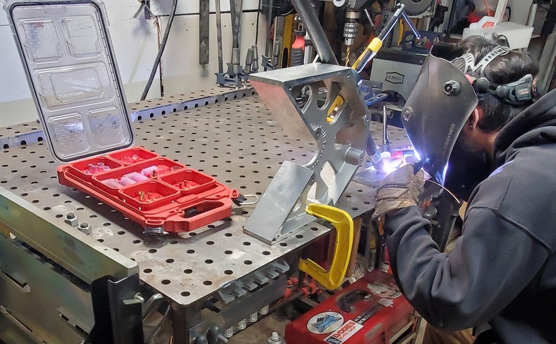 Może przedstawiać: Spawacz w czarnym swetrze z kapturem i masce spawalniczej pracuje nad metalowym projektem. Projekt jest zamocowany do metalowego stołu warsztatowego za pomocą ż&oacute;łtej klamry. Na stole warsztatowym znajduje się czerwona skrzynka narzędziowa z przezroczystą pokrywą.