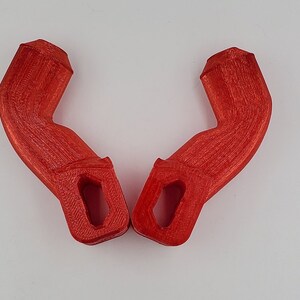 Puede incluir: Dos asas de pl&aacute;stico rojo impresas en 3D con forma curva y un agujero rectangular en el centro.