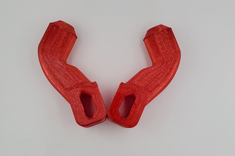 Puede incluir: Dos asas de pl&aacute;stico rojo impresas en 3D con forma curva y un agujero rectangular en el centro.