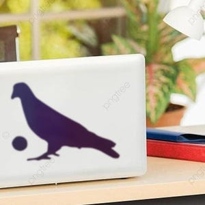 May include: A white laptop with a dark purple silhouette of a pigeon and a ball. The laptop is on a light-colored desk with a desk lamp, a red and blue folder, and a plant in the background. The laptop is open.