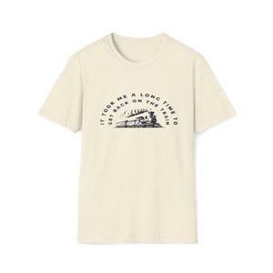 May include: A beige t-shirt with a vintage-style illustration of a steam train and the text "It took me a long time to get back on the train."