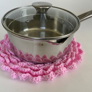 May include: A stainless steel saucepan with a glass lid rests on a handmade pink crochet trivet. The trivet has a ruffled edge and is designed to protect surfaces from heat. The kitchenware is set against a white background.