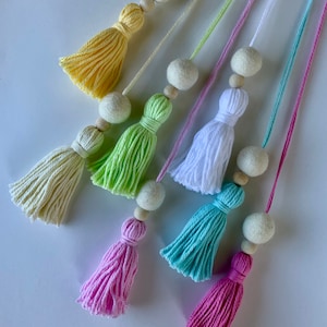 May include: A collection of colorful tassels in shades of yellow, green, white, blue, and pink. Each tassel is paired with a felt ball and wooden bead, strung on a matching cord. The tassels are arranged diagonally against a white background.