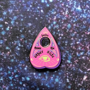 May include: A pink and purple planchette pin with the text "hello from the other side" and an eye symbol. The pin has a hole in the center and a purple border. The background is a dark blue fabric with white and blue specks.