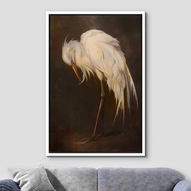 May include: A framed artwork depicting a white egret with its head bowed, highlighting the intricate details of its feathers. The bird is positioned on one leg against a dark brown backdrop, creating a contrast. The artwork is presented in a white frame.