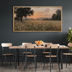 Puede incluir: Un cuadro de paisaje rectangular en un marco de madera clara que representa una puesta de sol sobre un campo de flores silvestres blancas y un gran &aacute;rbol. La escena presenta un cielo naranja suave, colinas lejanas y un ambiente oscuro.
