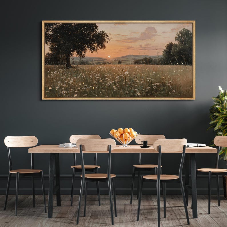 Puede incluir: Un cuadro de paisaje rectangular en un marco de madera clara que representa una puesta de sol sobre un campo de flores silvestres blancas y un gran &aacute;rbol. La escena presenta un cielo naranja suave, colinas lejanas y un ambiente oscuro.