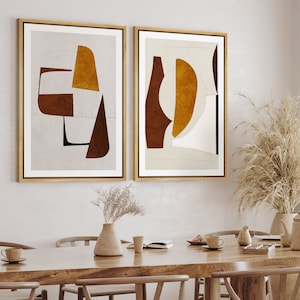 May include: Two framed abstract art prints with geometric shapes in brown, beige, and gold tones. The prints are hanging on a white wall above a wooden table.