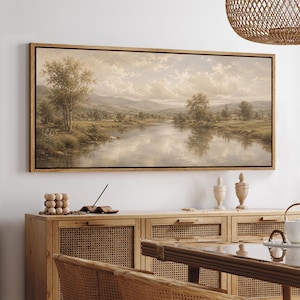 May include: A rectangular landscape painting in a thin dark frame depicting a calm river flowing through a valley with rolling hills, scattered trees, and a cloudy sky. The artwork features a muted, sepia-toned color palette with soft light.