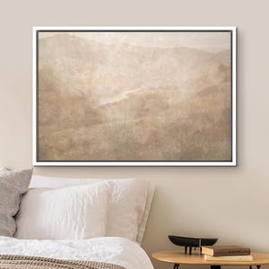 May include: A framed landscape art print featuring a muted, sepia-toned mountain range and a winding river. The artwork is encased in a white frame, creating a serene and natural aesthetic. The print is displayed above a bed with white pillows.