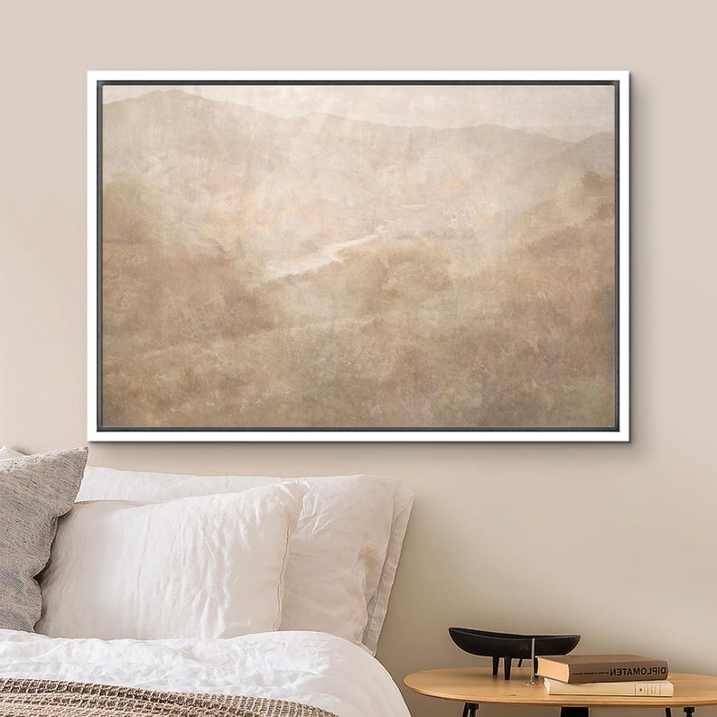 May include: A framed landscape art print featuring a muted, sepia-toned mountain range and a winding river. The artwork is encased in a white frame, creating a serene and natural aesthetic. The print is displayed above a bed with white pillows.