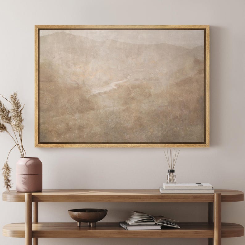 May include: A framed landscape art print featuring a muted color palette of beige, brown, and green. The artwork depicts a mountain range and a river. The frame is made of light-colored wood. The artwork is displayed on a wooden console table.