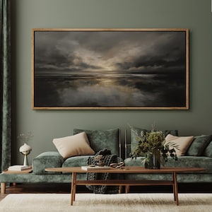 May include: A framed landscape painting with a dark, moody sky reflected in the water below. The artwork is in a wooden frame and hangs above a green velvet sofa with beige pillows. The room has a modern, minimalist aesthetic.