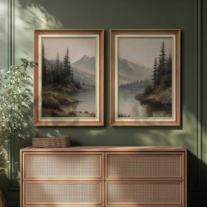 May include: Two framed landscape paintings depict a serene lake scene with mountains and evergreen trees. The artwork features a muted color palette with brown wooden frames and gold inner trim. The paintings are displayed above a wooden cabinet.