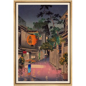 May include: A woodblock print depicting a rainy night scene in a Japanese city. The print shows a narrow street lined with traditional wooden buildings, with a woman walking under an umbrella in the foreground. The street is wet from the rain, and the buildings are illuminated by lanterns. The print is framed in a wooden frame.