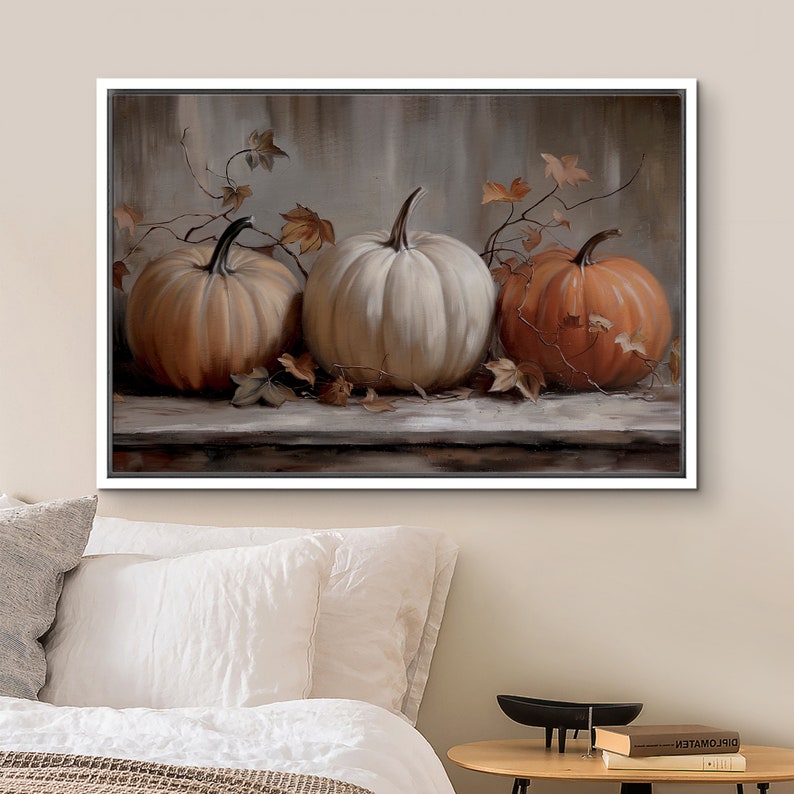 May include: A framed canvas art print featuring three pumpkins in shades of orange and white. The pumpkins are arranged on a wooden surface with autumn leaves and vines. The background is a muted, neutral color.