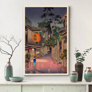 May include: A vintage Japanese woodblock print depicting a narrow street scene at night. The street is wet from rain and lit by lanterns. A single figure walks down the street, carrying an umbrella. The print is framed in a light wood frame.
