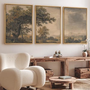 May include: Three framed landscape paintings with a rustic, vintage aesthetic. The paintings depict trees, fields, and a cloudy sky. The paintings are hung on a white wall above a wooden table and a white armchair.