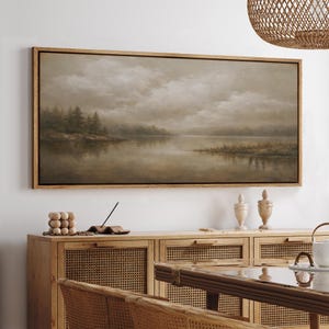 May include: A framed landscape painting depicts a serene lake scene under a cloudy sky. The artwork features muted tones of brown, gray, and beige, with a wooden frame. The painting is displayed above a wooden cabinet with woven rattan doors.
