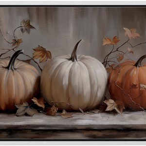 May include: A framed painting featuring three pumpkins in shades of orange and white. The pumpkins are arranged on a wooden surface, with dried leaves and vines adding to the autumn theme. The background is a muted gray.