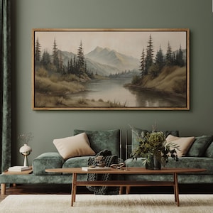 May include: A framed landscape painting depicts a serene mountain scene with a lake and evergreen trees. The artwork is in a wooden frame and hangs above a green velvet sofa with beige pillows. The room has a neutral color palette.