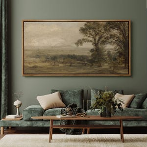 May include: A framed landscape painting with a muted color palette hangs above a green velvet sofa. The artwork depicts a rural scene with trees, fields, and a distant horizon. The frame is a warm, natural wood tone.