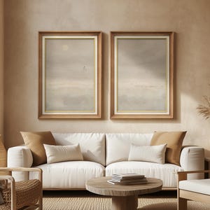 May include: Two framed landscape art prints with a neutral color palette. The artwork features a soft, muted color scheme with a light brown wooden frame and a gold inner border. The prints are displayed above a white sofa with neutral-colored pillows.