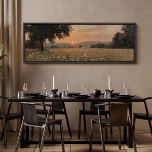Puede incluir: Un cuadro de paisaje rectangular en un marco negro que representa una puesta de sol sobre un campo de flores silvestres blancas y un &aacute;rbol grande. La escena muestra colinas bajo un cielo naranja suave con peque&ntilde;as part&iacute;culas luminosas.