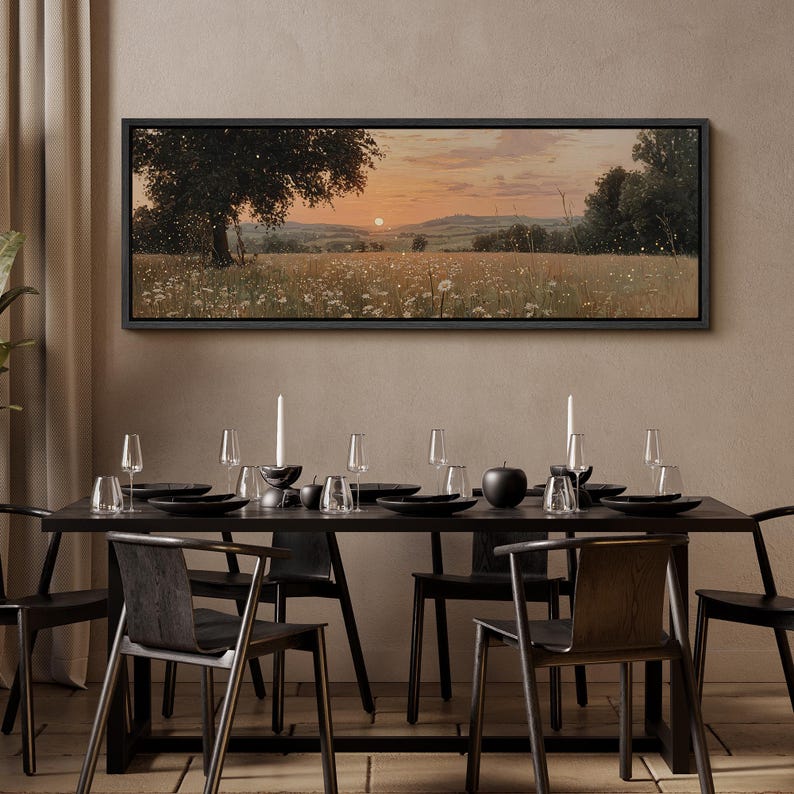 Puede incluir: Un cuadro de paisaje rectangular en un marco negro que representa una puesta de sol sobre un campo de flores silvestres blancas y un &aacute;rbol grande. La escena muestra colinas bajo un cielo naranja suave con peque&ntilde;as part&iacute;culas luminosas.