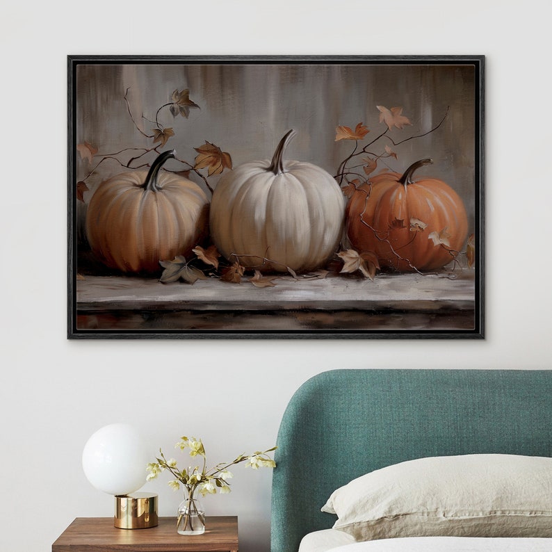 May include: A framed canvas art print featuring three pumpkins in shades of orange and white. The pumpkins are arranged on a wooden surface with autumn leaves and vines. The artwork has a neutral background and a black frame.