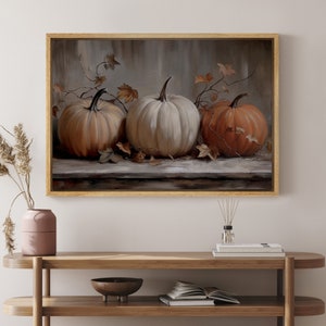 May include: A framed artwork featuring three pumpkins in shades of orange and white. The pumpkins are arranged on a wooden surface with autumn leaves and vines. The artwork is in a gold frame, suitable for fall decor.