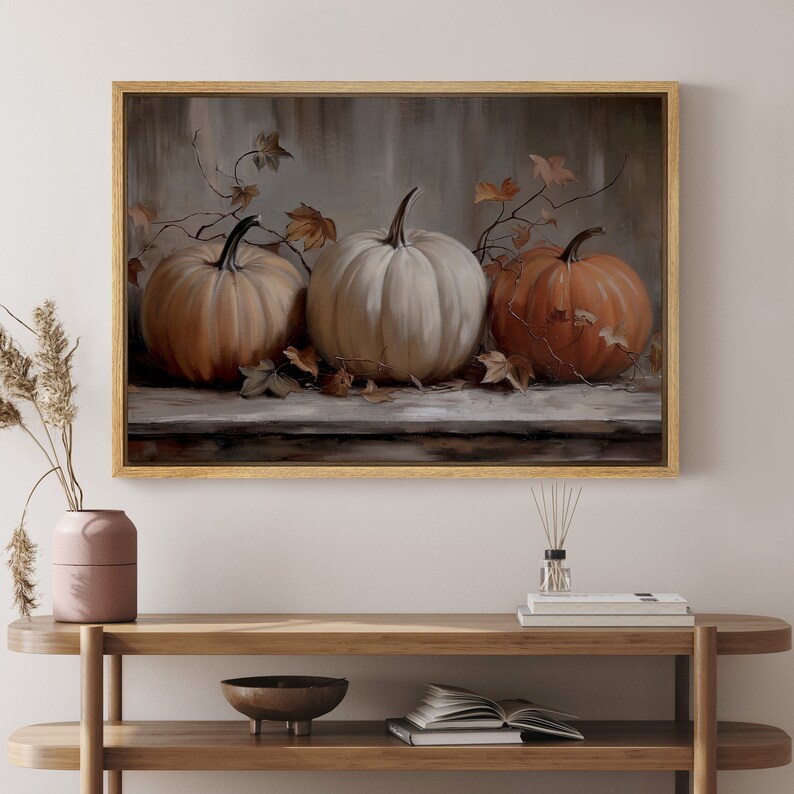 May include: A framed artwork featuring three pumpkins in shades of orange and white. The pumpkins are arranged on a wooden surface with autumn leaves and vines. The artwork is in a gold frame, suitable for fall decor.