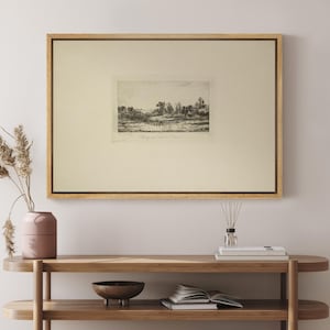 May include: A framed black and white landscape etching. The artwork is displayed in a light wood frame with a cream-colored mat. The etching depicts a rural scene with trees and a body of water. The artwork is displayed on a wooden console table.