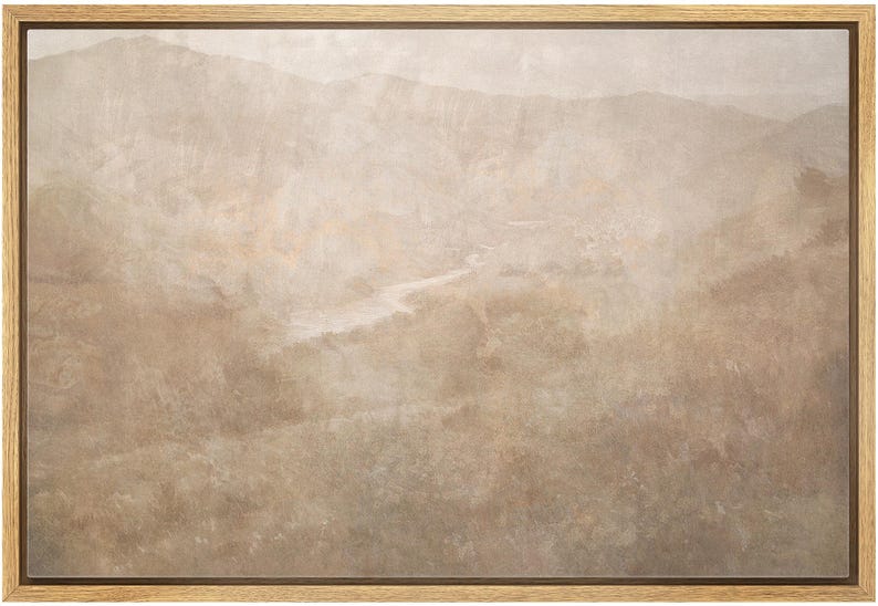 May include: A framed landscape art print featuring a muted color palette of beige, tan, and brown. The artwork depicts a mountainous terrain with a river winding through the valley. The frame is made of light-colored wood.