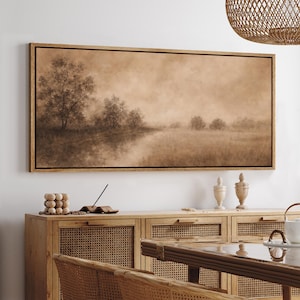 May include: A sepia-toned landscape painting in a wooden frame. The artwork depicts a serene scene of trees reflected in a body of water, with a hazy atmosphere. The painting is displayed above a wooden cabinet with woven panels.