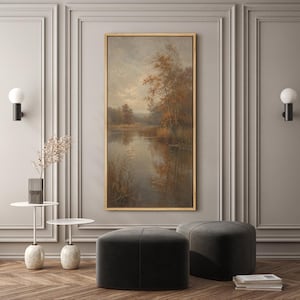 May include: A vertical rectangular framed wall art piece depicting a landscape painting of a calm lake surrounded by autumn trees in shades of brown, orange, and muted green. The artwork is enclosed in a thin gold frame and mounted on a wall.