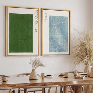 May include: Two framed art prints with a Japanese aesthetic. One features a green field with flying birds, the other a blue and white scene. Both have gold frames and Japanese text. The prints are displayed above a wooden table.